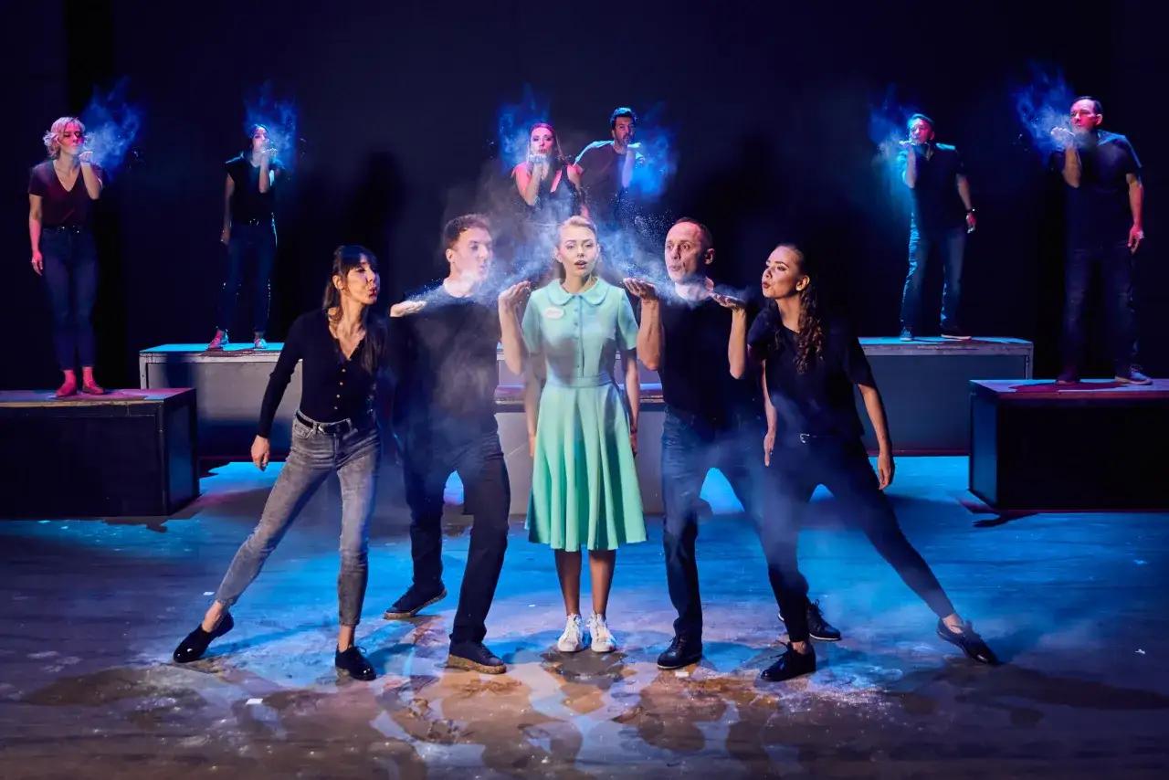 Aktorzy w teatrze Roma, w tym waitress, dmuchają w niebieski proszek, tworząc efektowne chmury.