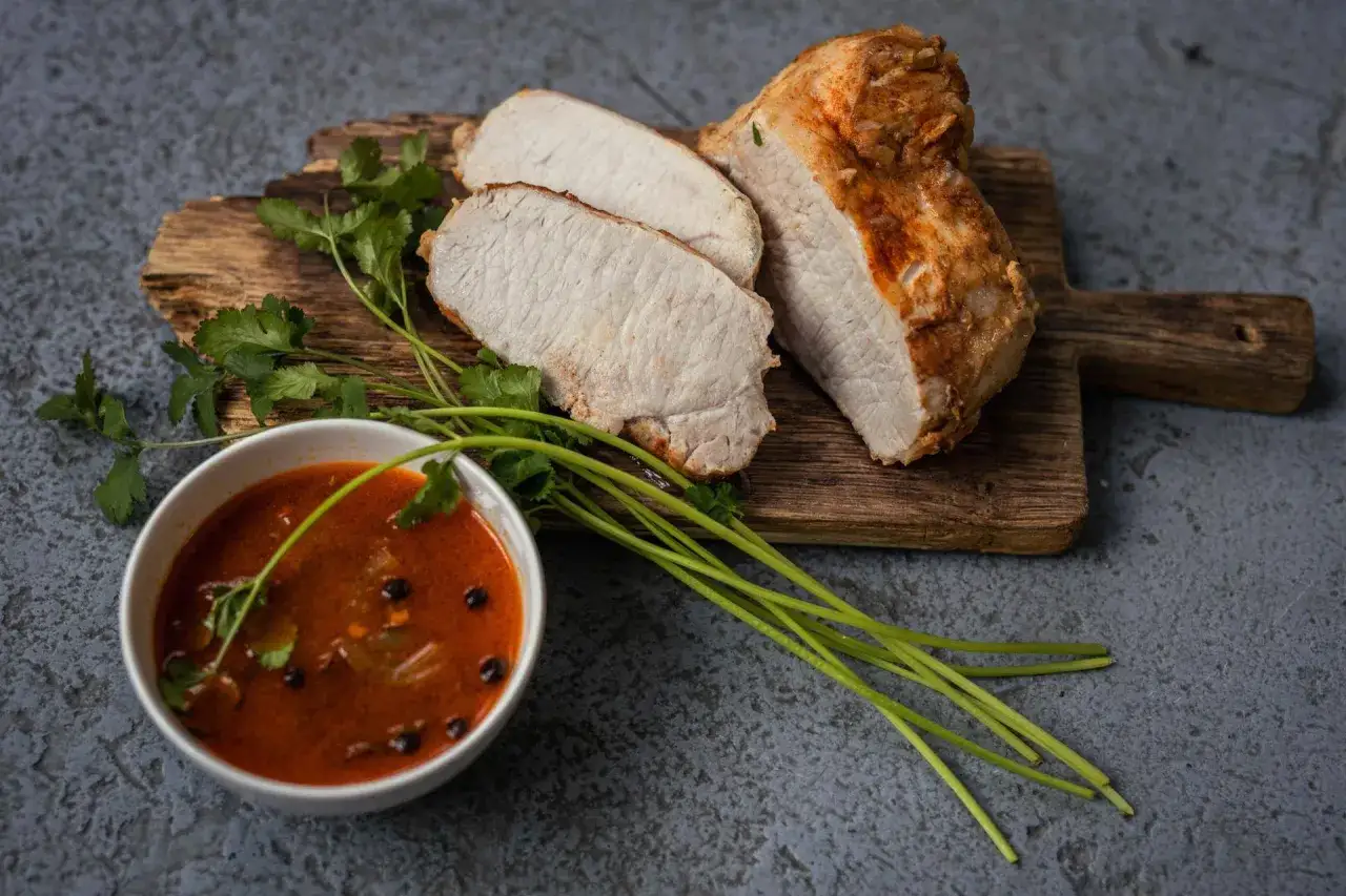 Kawałek pieczonego schabu, pokrojony na plastry, z miseczką gulaszu i gałązkami kolendry. Idealny pomysł, ile dusić schab do miękkości.