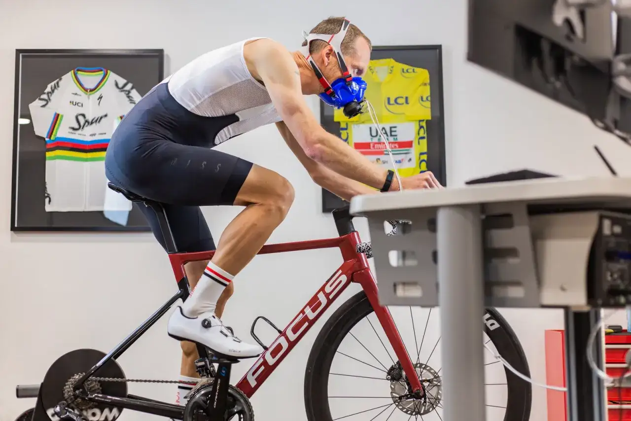 Kolarz w masce mierzy VO2 max na rowerze stacjonarnym, obok wiszą koszulki kolarskie.