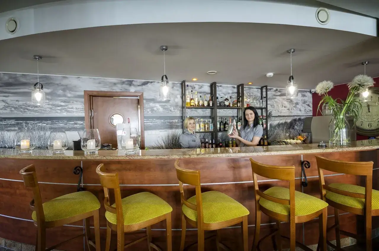 Bar w hotelu La Siesta, gdzie dwie barmanki przygotowują drinki. W tle półki z alkoholem i dekoracja z motywem plażowym.