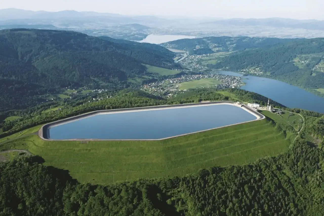 Zbiornik szczytowy na górze Żar, otoczony zielonymi wzgórzami i lasami, z widokiem na jezioro i miasteczko w oddali.