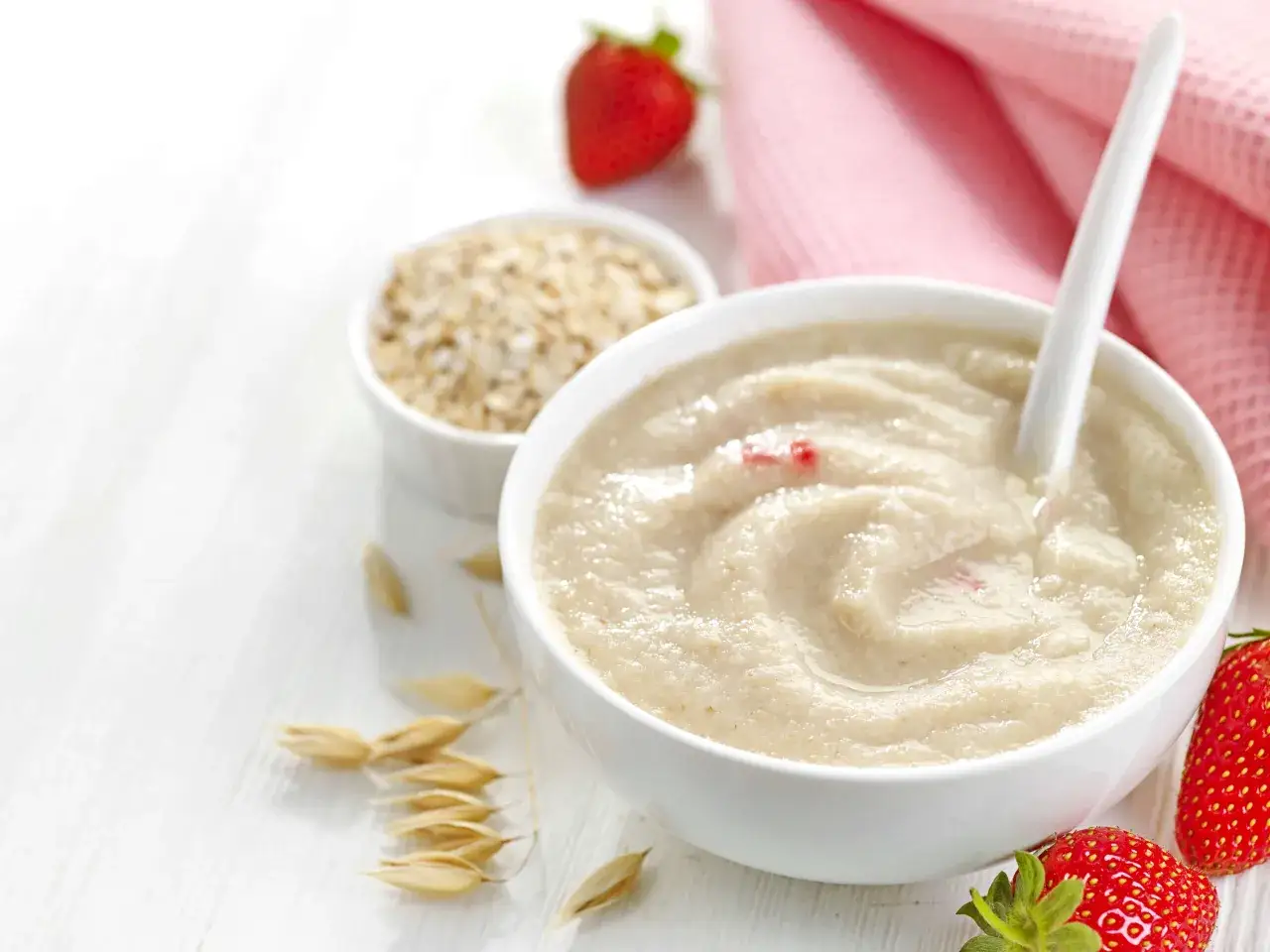 Owsianka z truskawkami, idealna dla maluch&oacute;w po 4 miesiącu. Obok miseczki z kaszką leżą płatki owsiane i świeże owoce.