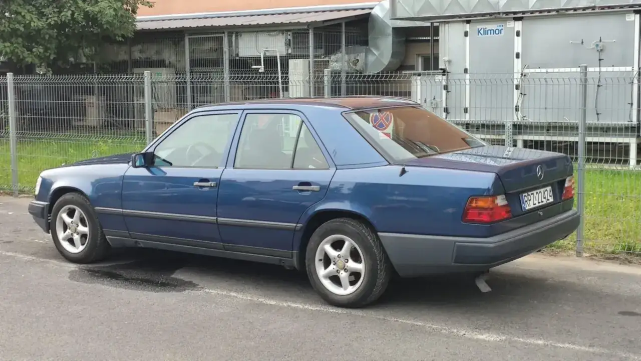 Niebieski Mercedes W124 z lat 90. stoi zaparkowany przy płocie.