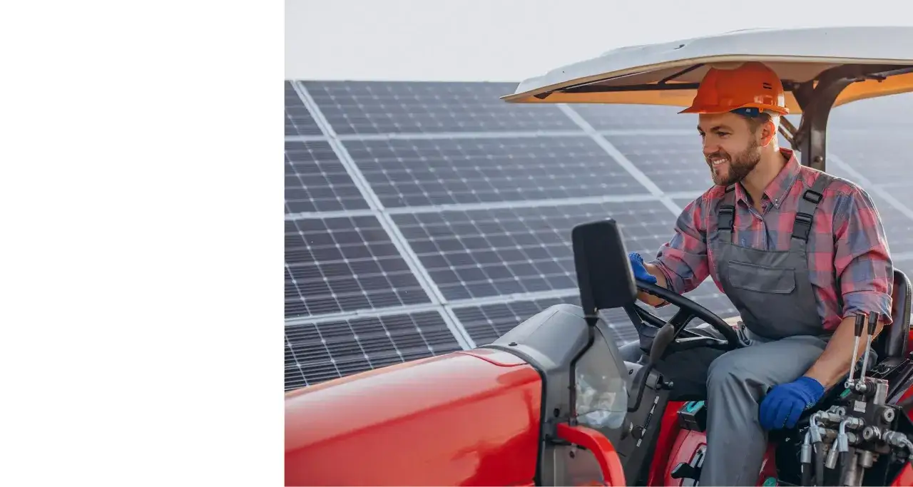 Uśmiechnięty rolnik w kombinezonie i kasku siedzi za kierownicą traktora na tle paneli fotowoltaicznych. Nowoczesne rozwiązania dla gospodarstwa.
