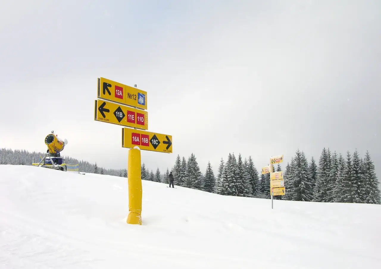 Żółte tablice z kolorami tras narciarskich wskazują drogę. W tle śnieżny stok, las i armatka śnieżna.
