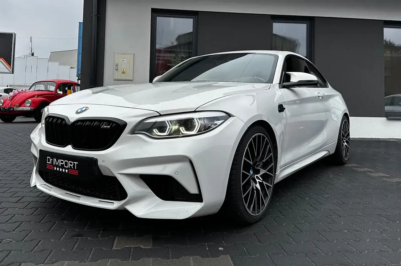 Białe BMW M2 z czarnym grillem i felgami. Obok czerwony klasyk. Sprawdź aktualną bmw m2 cena!