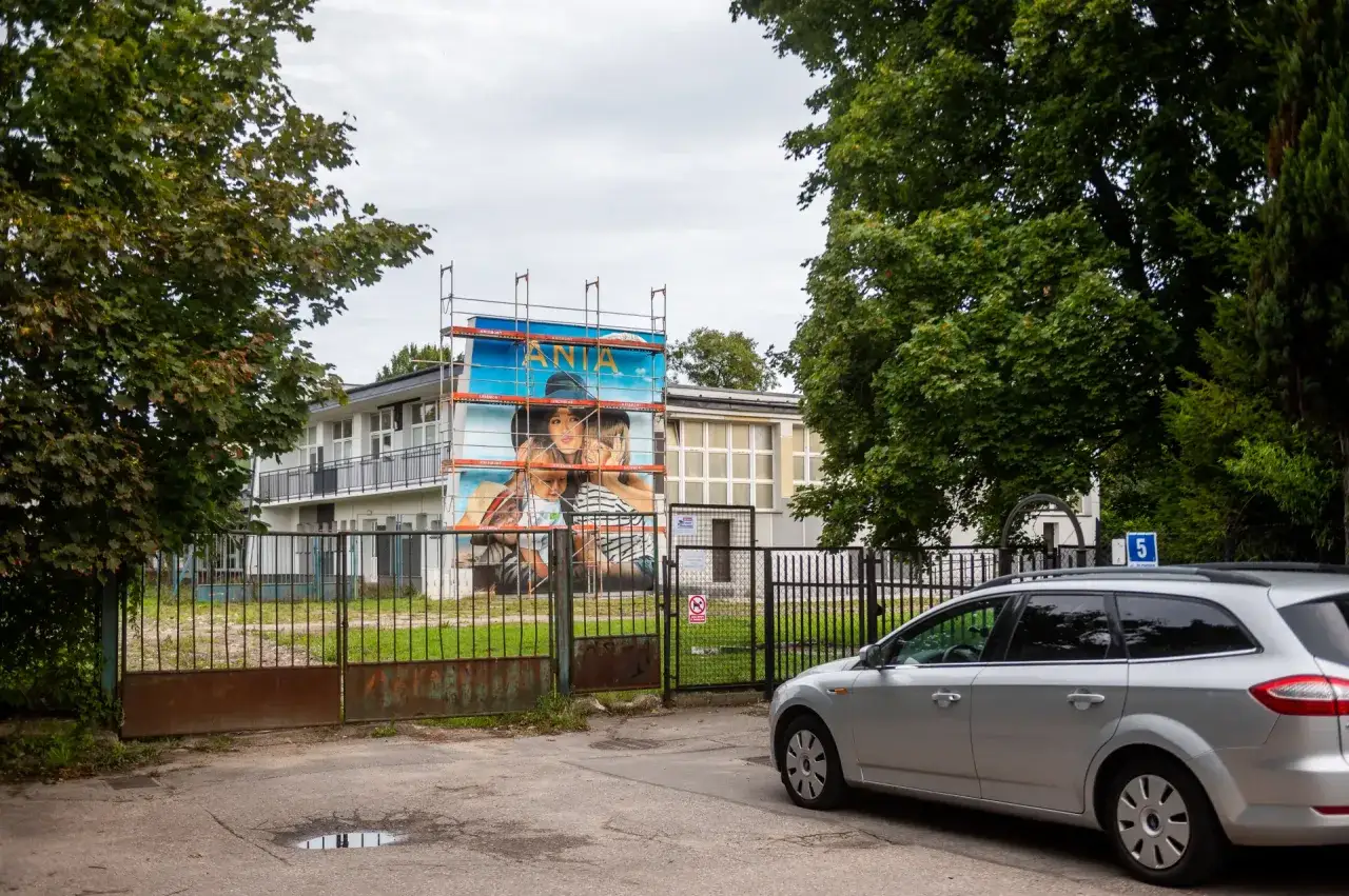Wielki mural z Anną Przybylską i dziećmi na budynku. Obok brama i srebrne auto. To miejsce upamiętnia jej dziedzictwo, choć jej wykształcenie nie jest tu widoczne.