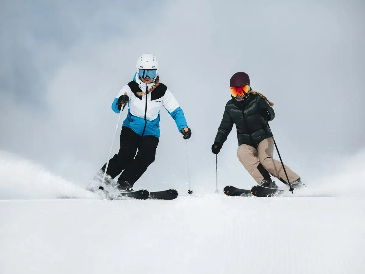 Dwie kobiety na nartach damskich zjeżdżają po ośnieżonym stoku, tworząc tumany śniegu.