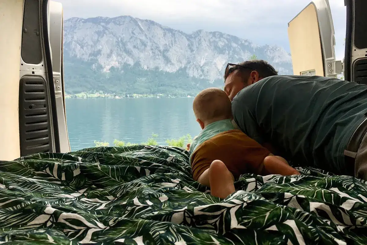 Ojciec i dziecko leżą na pościeli w liście, patrząc na jezioro i góry z wnętrza kampera.