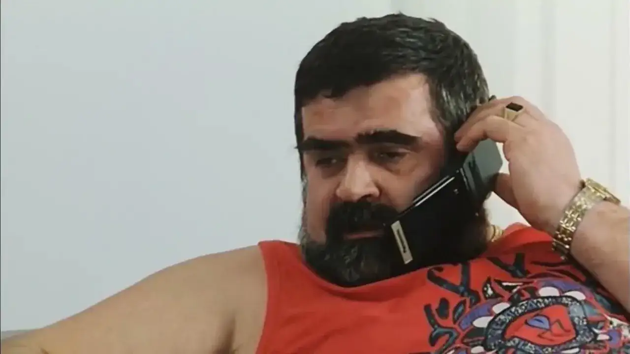 Gruby gangster z filmu Kiler, z bujną brodą i w czerwonej koszulce, rozmawia przez telefon.