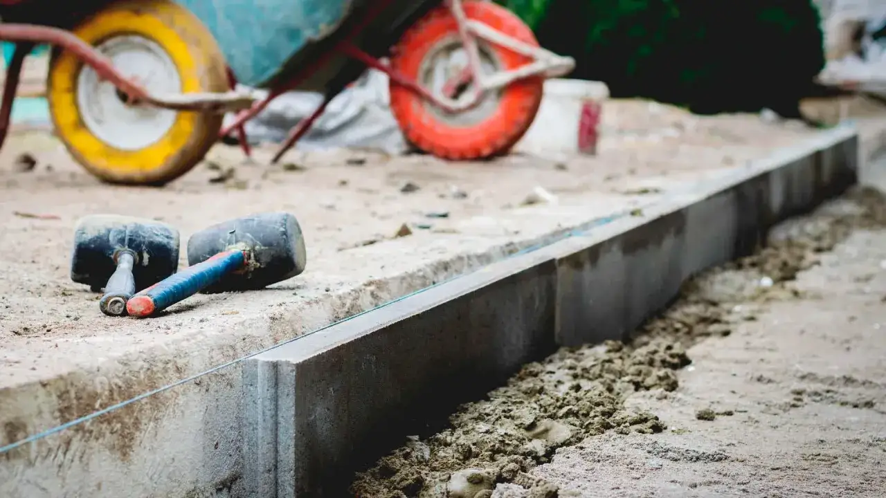 Dwa gumowe młotki leżą obok świeżo ułożonych krawężników. W tle widać taczkę, sugerującą prace budowlane i potrzebę odpowiedniej podsypki pod krawężniki proporcje.