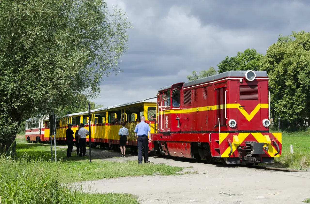 Czerwona lokomotywa i żółte wagony na szlaku kolejki wąskotorowej. Ludzie wsiadają do pociągu.