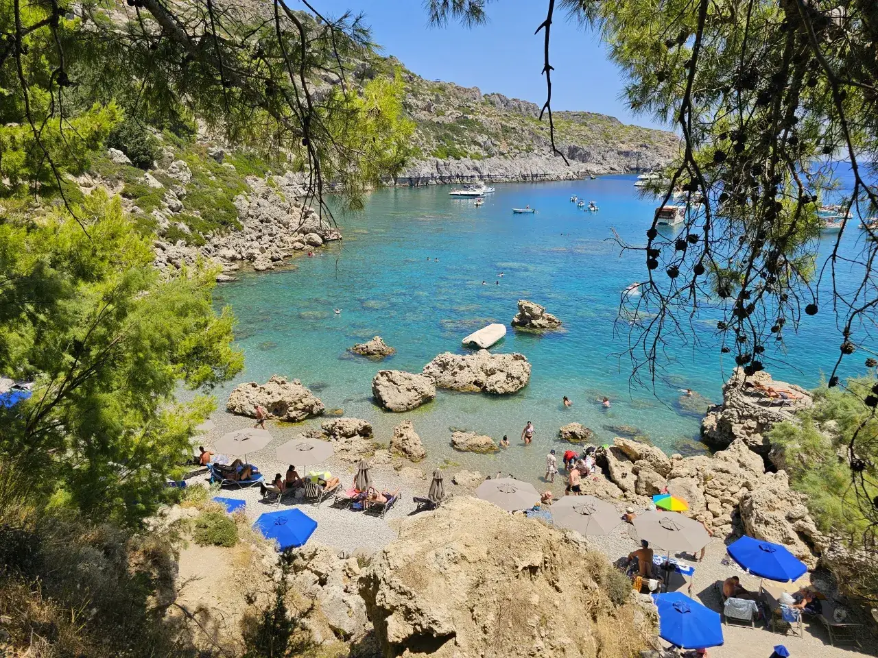 Słoneczna zatoka z turkusową wodą, kamienistą plażą i ludźmi odpoczywającymi pod parasolami. Idealne miejsce, by poczuć, kiedy najlepiej jechać do Grecji.