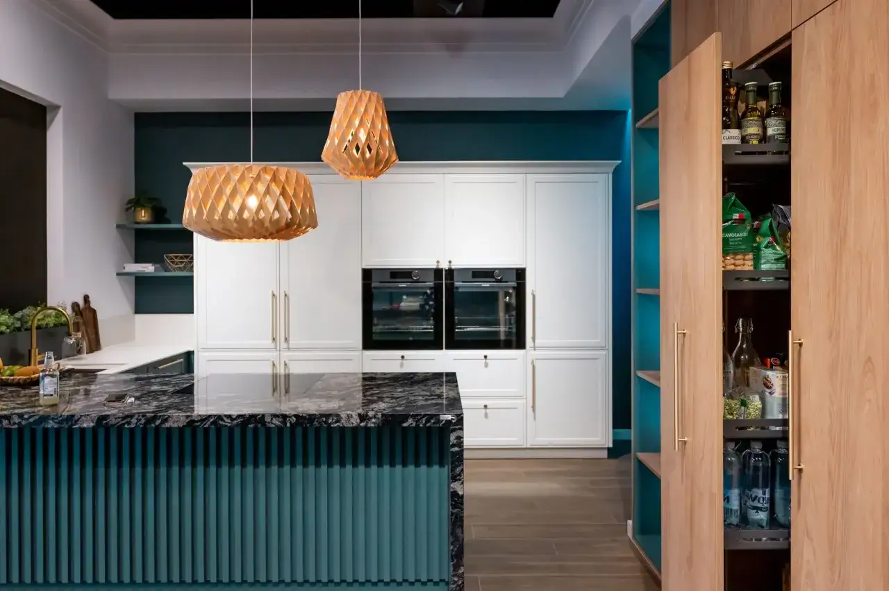 Elegancka kuchnia modern classic z wyspą i drewnianymi lampami. Białe fronty szafek, wbudowane piekarniki i praktyczna spiżarnia.