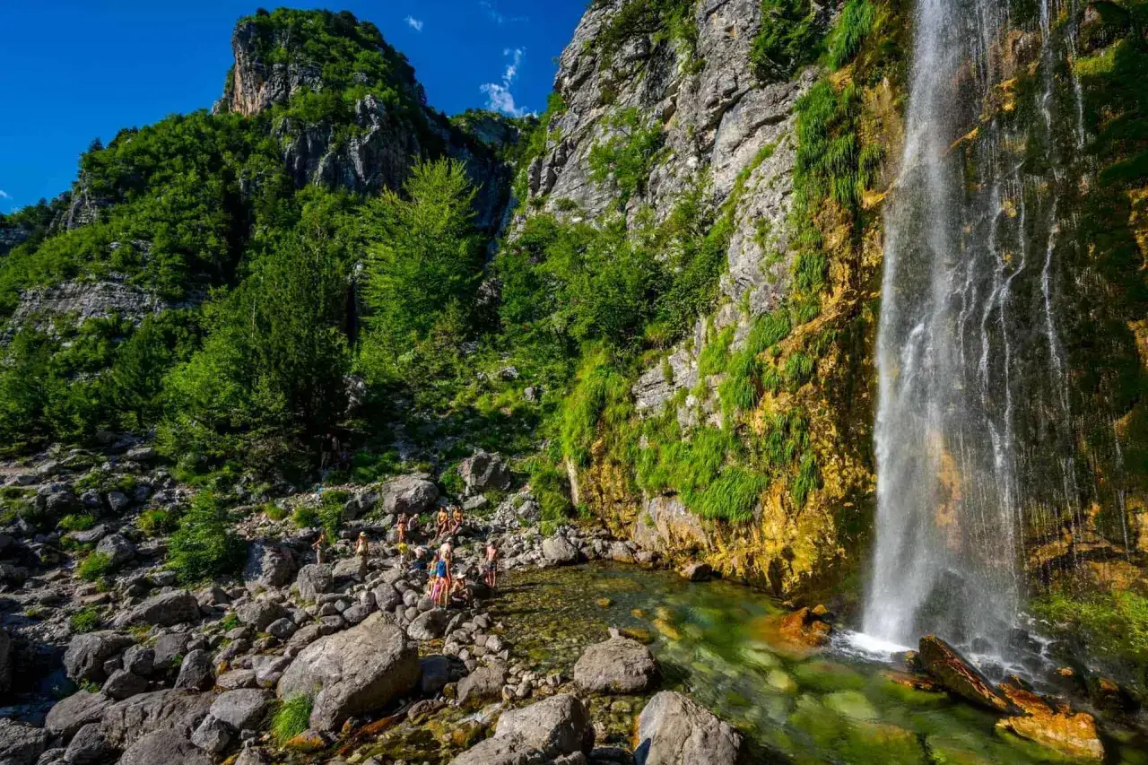 Albania wodospady: Grupa ludzi podziwia kaskadę spadającą po skalnej ścianie porośniętej zielenią, w krystalicznie czystym basenie.