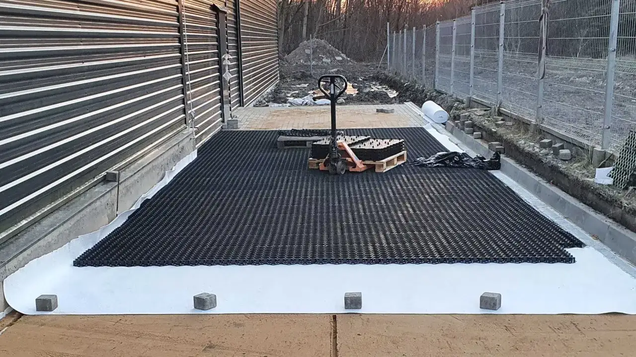 Układanie geosiatki na podwórku to tani sposób na utwardzenie terenu. Widać palety z materiałem i wózek widłowy.