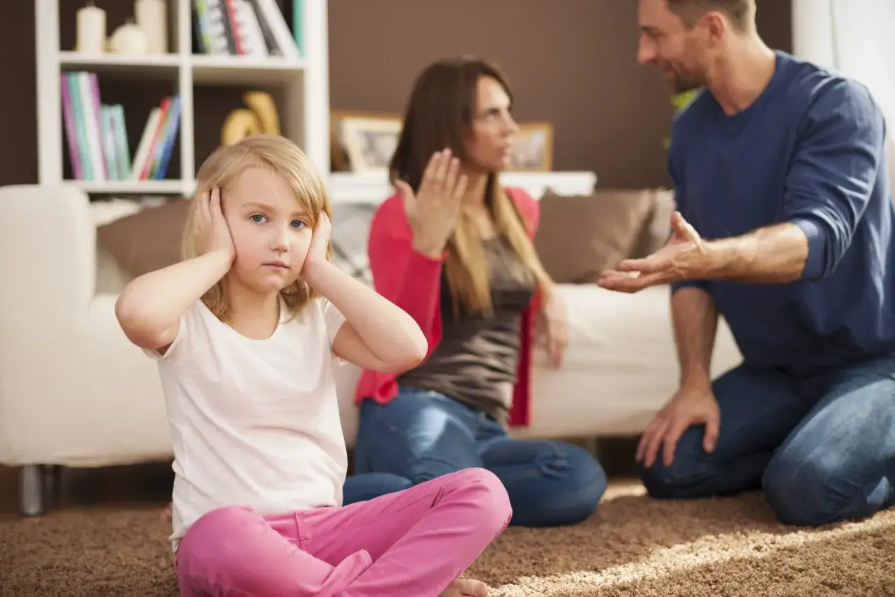 Dziewczynka zatyka uszy, pr&oacute;bując zagłuszyć kł&oacute;tnię rodzic&oacute;w. W tle mama i tata dyskutują, a ona czuje się zagubiona, jakby miała problemy w szkole i nie wiedziała, jak sobie z nimi poradzić.