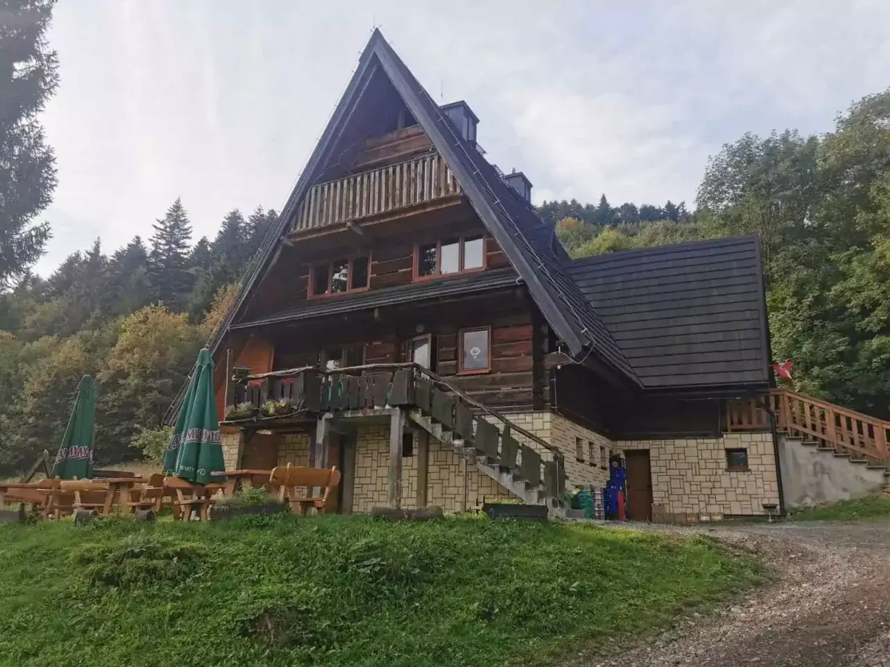 Drewniana bac&oacute;wka pod honem, otoczona zielenią i lasem, zaprasza do odpoczynku. Na zewnątrz stoliki i ławki czekają na gości.