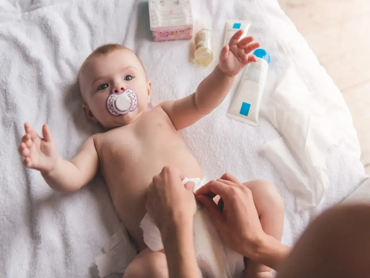 Rączki mamy zakładają pampersa chłopcu. Na ręczniku leżą kosmetyki i chusteczki.
