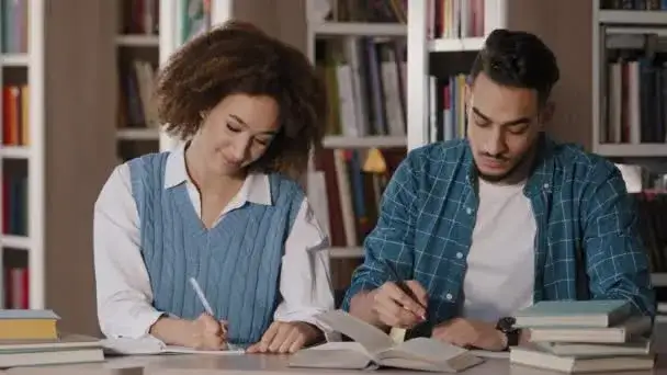 student w bibliotece, otoczony książkami, robi notatki