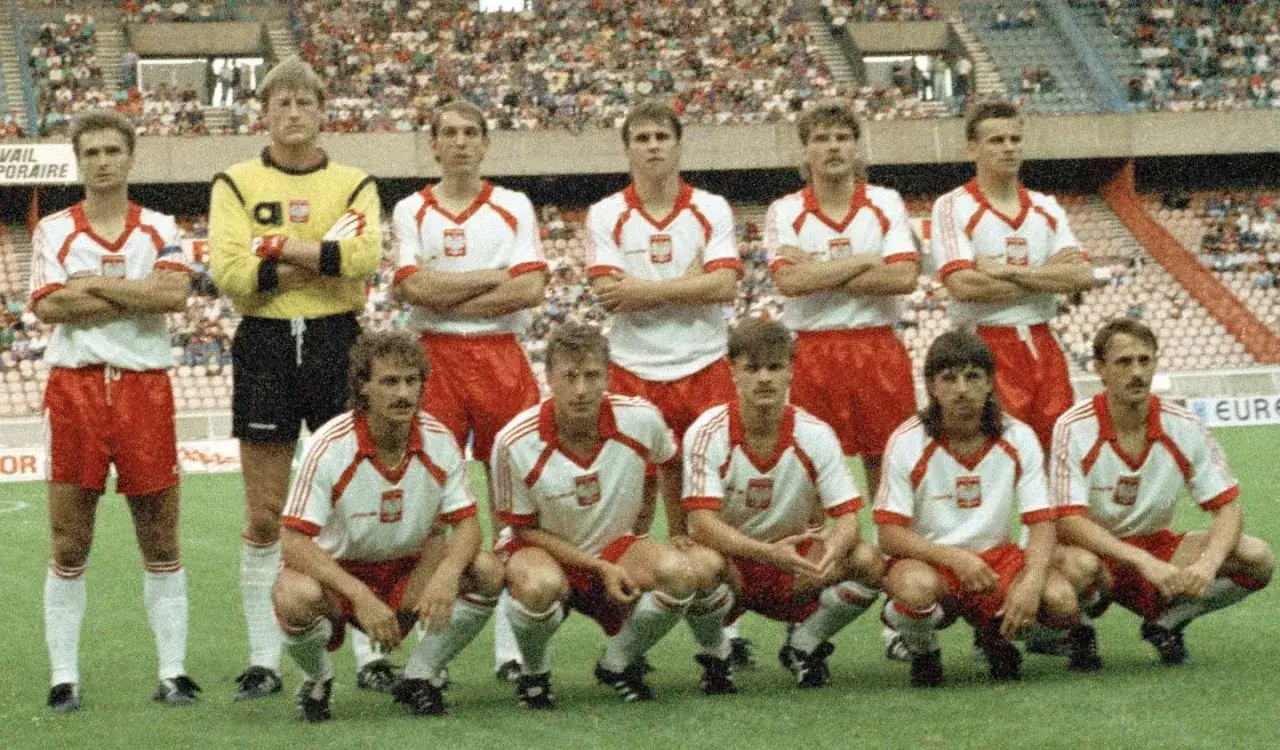 Drużyna polscy piłkarze lat 90. w białych koszulkach z czerwonymi pasami i czerwonych spodenkach, pozuje na boisku przed meczem.