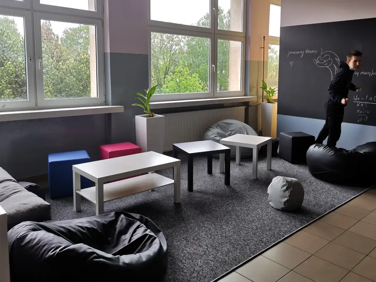 Przytulny kącik relaksu w szkole z pufami, stolikami i tablicą.