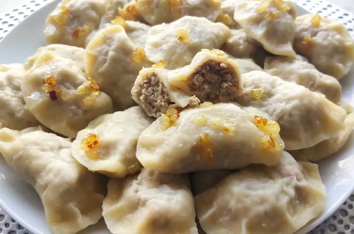 Talerz pełen pysznych, domowych pierog&oacute;w z mięsem i kapustą, posypanych skwarkami. Jeden pier&oacute;g przekrojony, ukazujący bogate nadzienie.