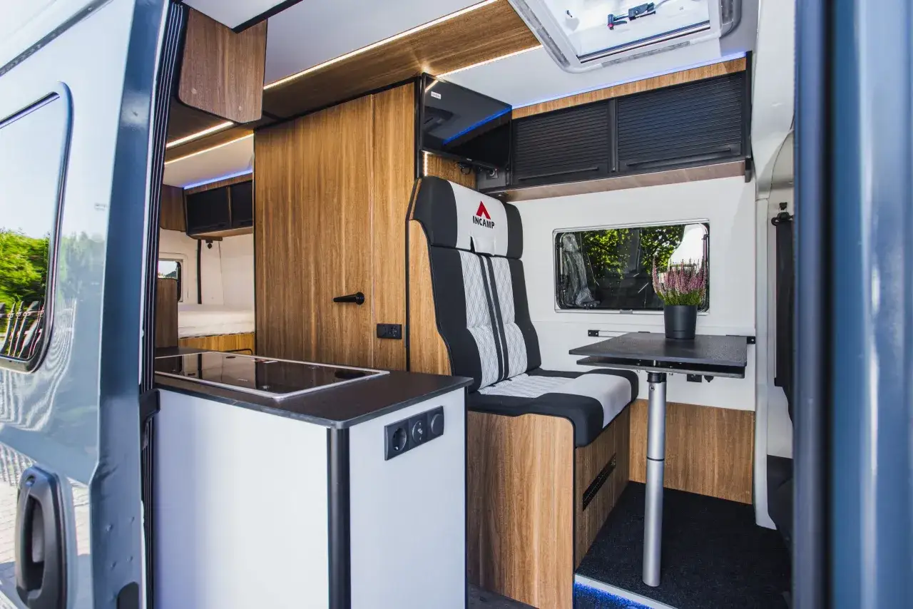 Nowoczesne wnętrze kampera z aneksem kuchennym, wygodnym fotelem i stolikiem. Drewniane wykończenia dodają przytulności.