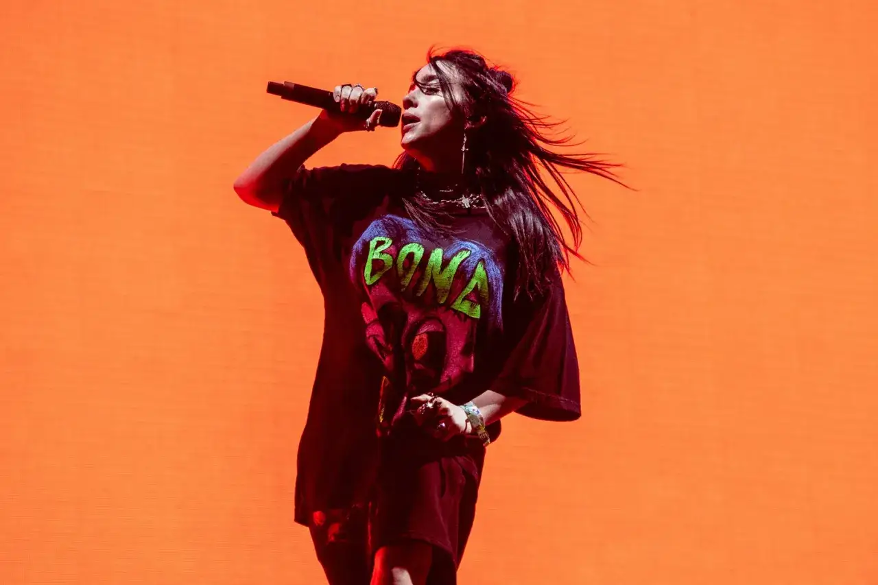 Piosenkarka Billie Eilish na scenie, z włosami rozwianymi przez wiatr, śpiewa do mikrofonu w czarnej koszulce z neonowym napisem.