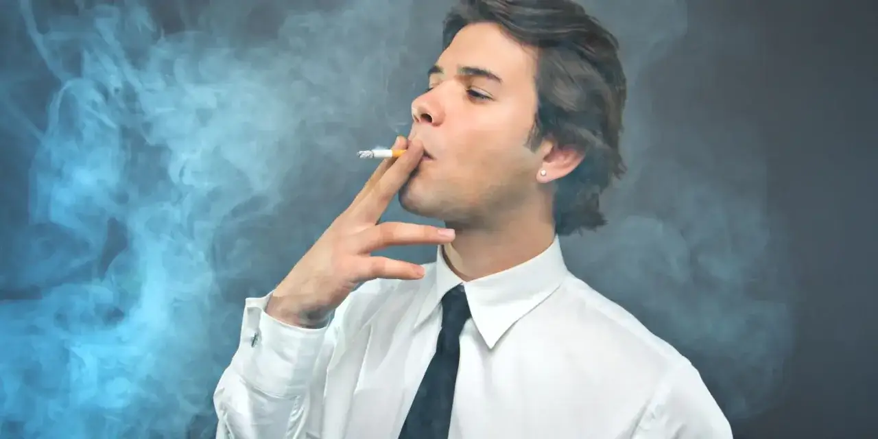 Młody mężczyzna w białej koszuli i krawacie pali papierosa, otoczony kłębami dymu. Czy kaszel palacza zawsze oznacza raka?