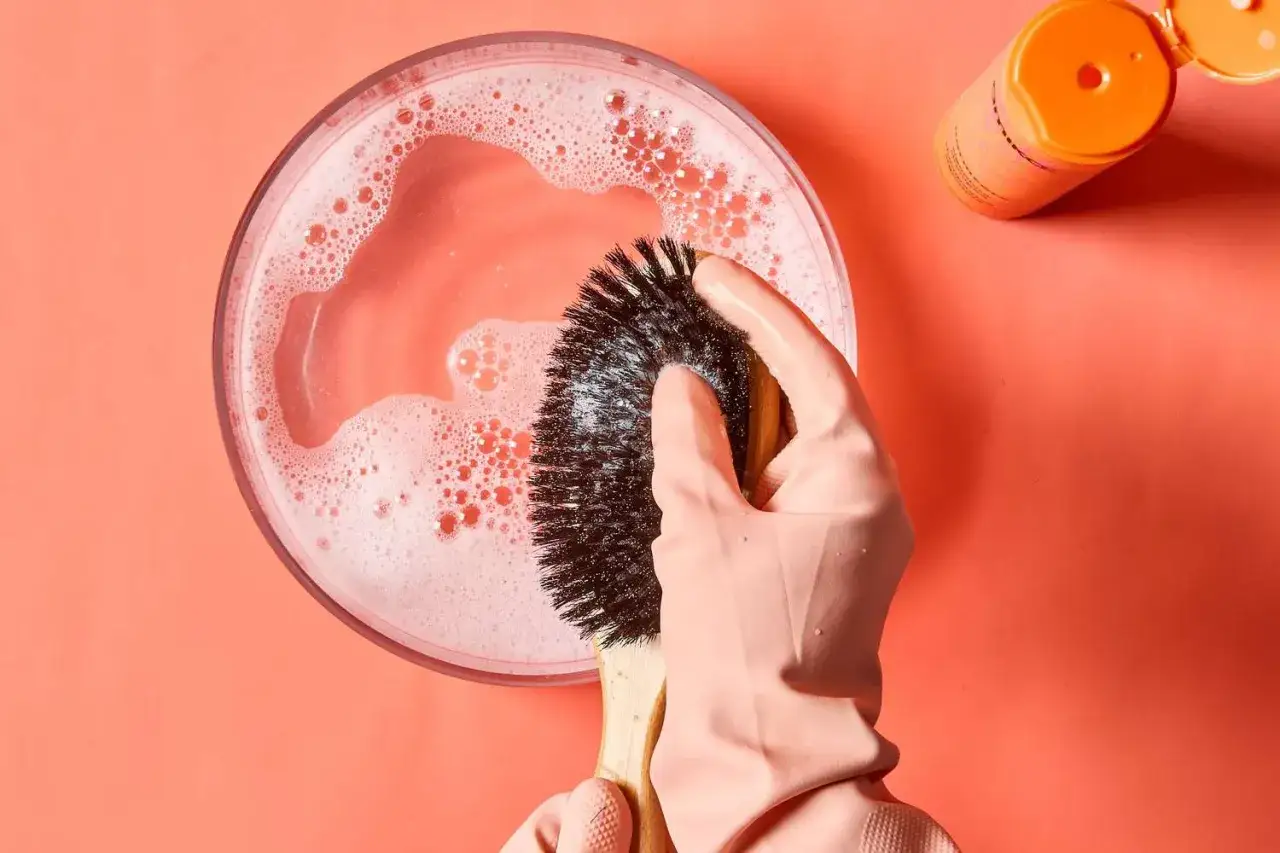 Rękawiczka w r&oacute;żowym kolorze czyści szczotkę do włos&oacute;w w wodzie z mydłem. Dowiedz się, jak wyczyścić szczotkę do włos&oacute;w.