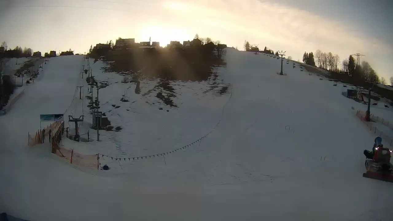 Ośnieżony stok narciarski na olczańskim wierchu z wyciągami i domkami w tle. Na dole pracuje ratrak.