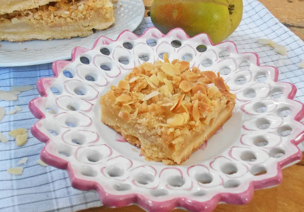 Pyszne ciasto z gruszkami, posypane płatkami migdałów, na ozdobnym talerzyku. Obok leży dojrzała gruszka.