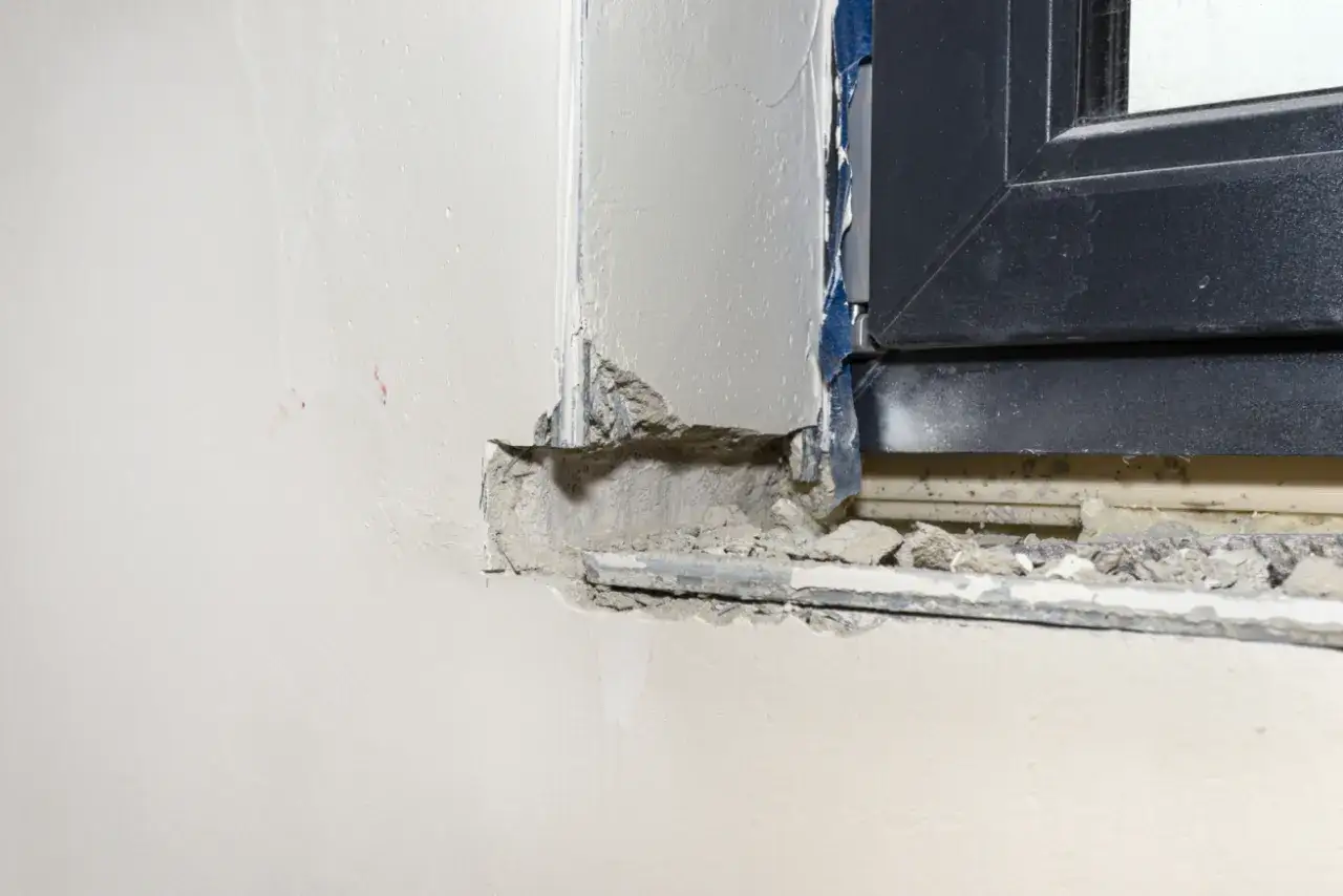 Fragment ściany z oknem i luźnym betonem. Czy parapet wewnętrzny musi być wpuszczony w ścianę? Widać resztki zaprawy i taśmę.