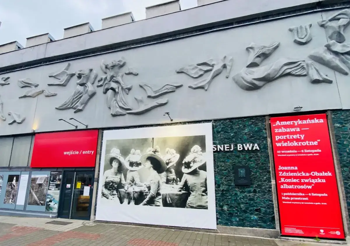 Galeria sztuki Katowice zaprasza na wystawy. Na fasadzie rzeźba i plakaty, w tym portrety kobiet w kapeluszach.