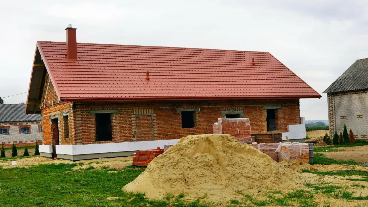 Budowa domu z czerwonej cegły, czerwony dach, komin, piasek i materiały budowlane. Wkr&oacute;tce ulga na remont.