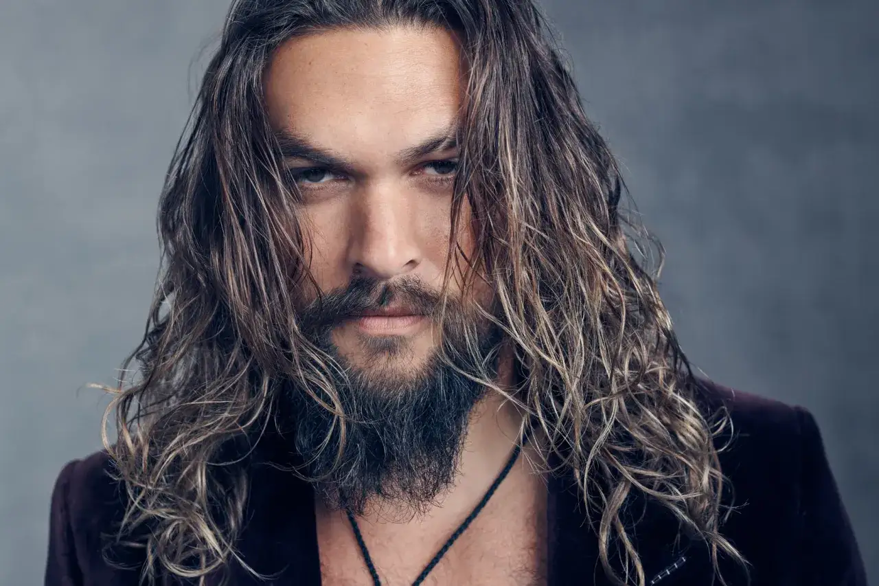 Jason Momoa, znany z film&oacute;w, seriali i program&oacute;w, z burzą włos&oacute;w i zarostem, w aksamitnej marynarce.