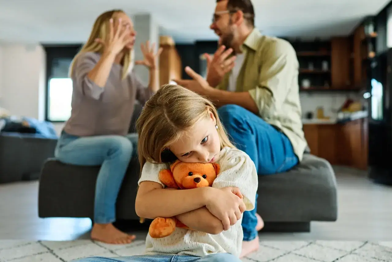 Smutna dziewczynka tuli misia, podczas gdy rodzice kłócą się w tle. To obraz trudnych chwil, gdy rozwód a dzieci stają się problemem.