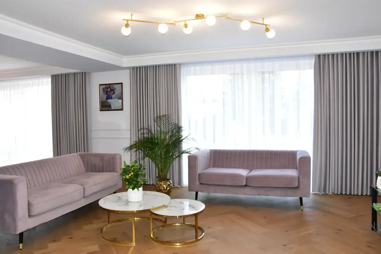 Eleganckie, szare firany na szynach tworzą przytulną atmosferę w salonie z różowymi sofami i marmurowymi stolikami.