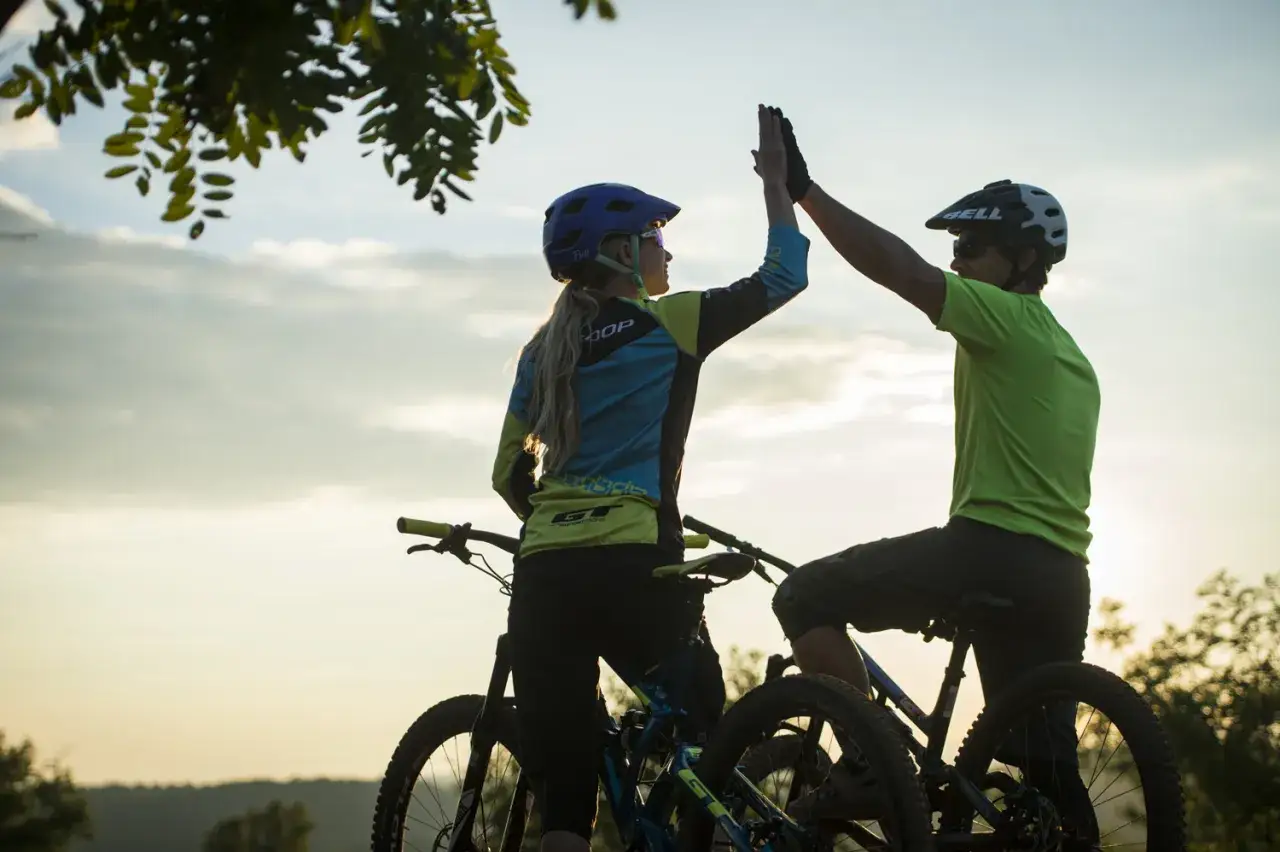 Dwóch rowerzystów w kaskach daje sobie piątkę na tle zachodzącego słońca. Cieszą się udanym przejazdem po torze rowerowym.