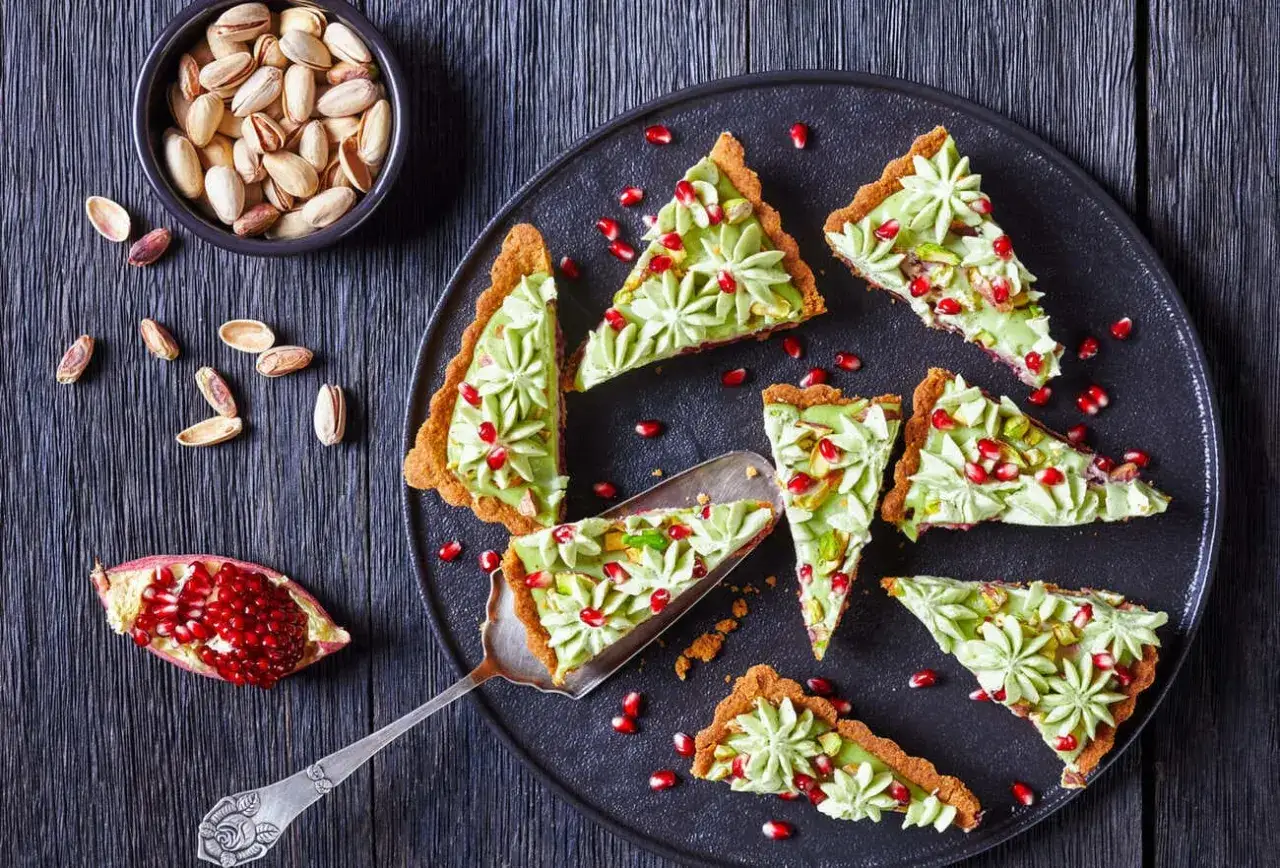 Kawałki tarty z kremem pistacjowym, ozdobione pestkami granatu i posiekanymi pistacjami. Obok miseczka z pistacjami i połówka granatu.