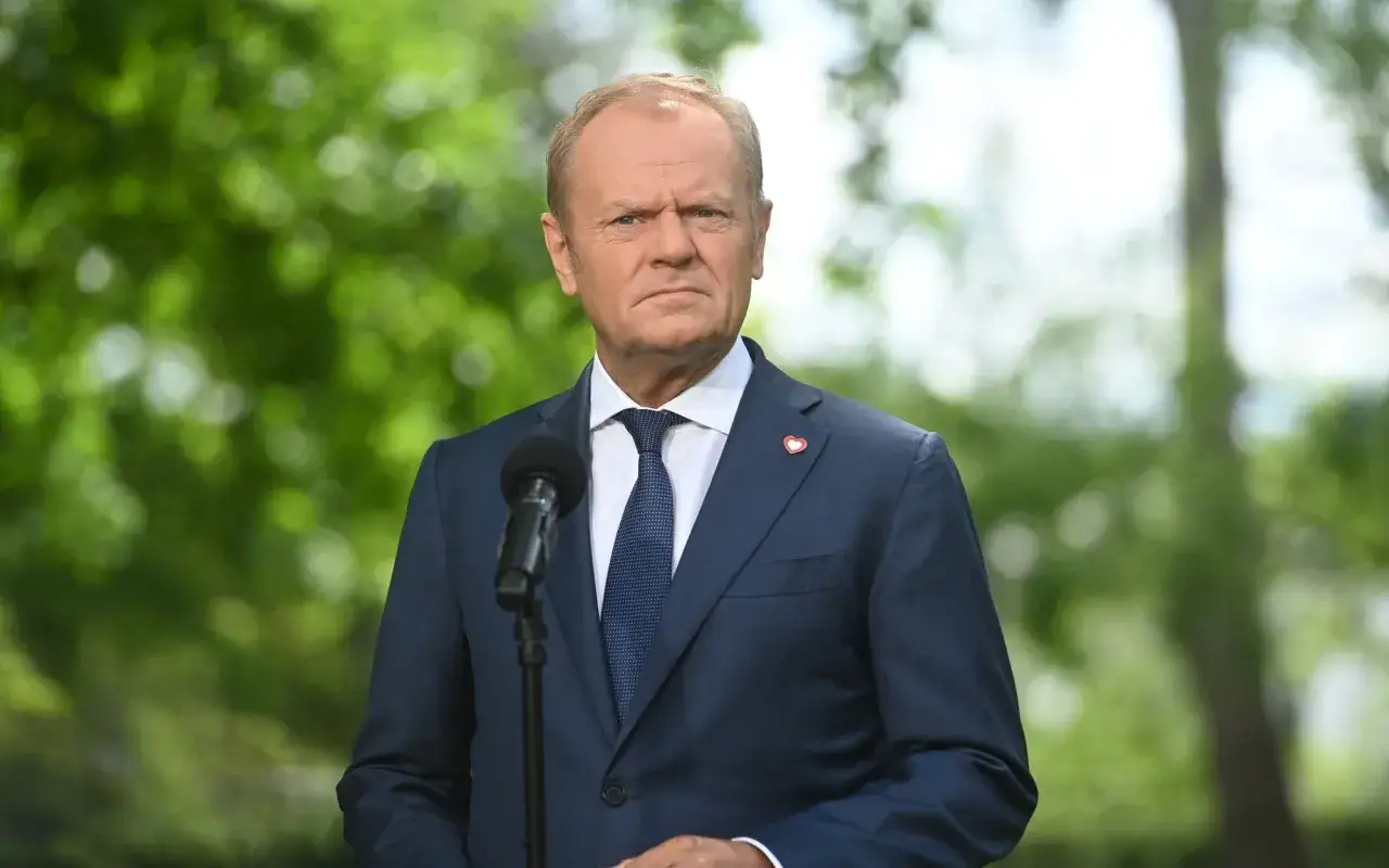 Donald Tusk w granatowym garniturze i krawacie, z serduszkiem na klapie, przemawia przy mikrofonie.