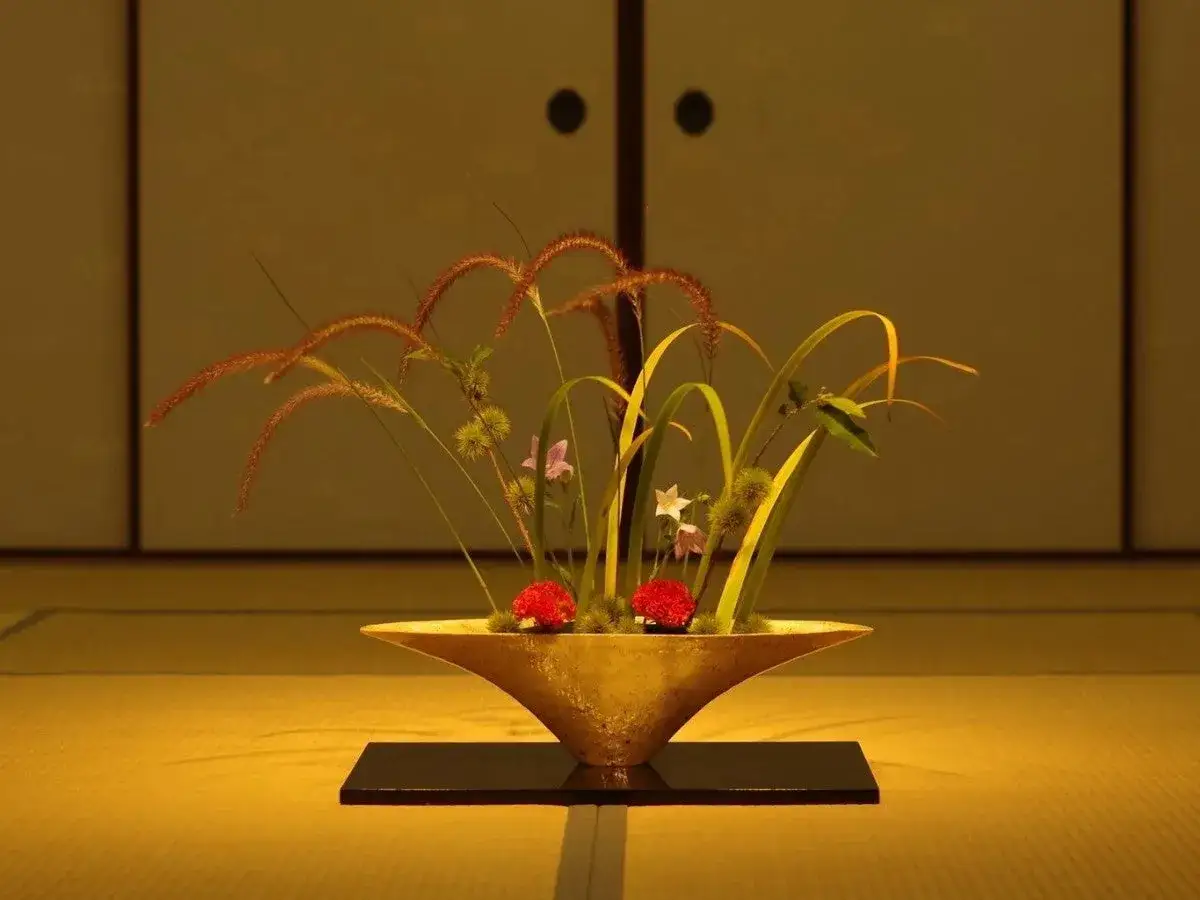Złota misa z trawami, liśćmi i dwoma czerwonymi kwiatami. Kompozycja ikebany kwiatowej w minimalistycznym wnętrzu.