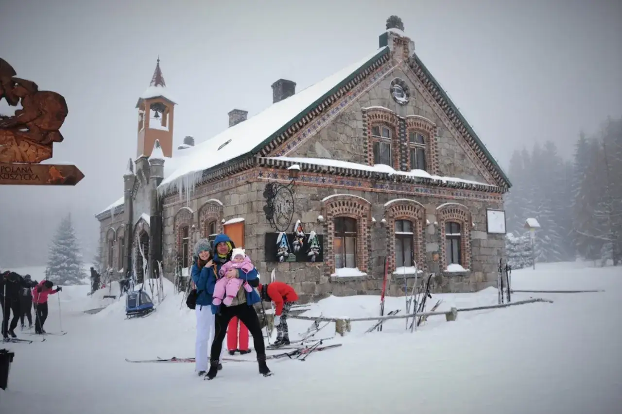 Rodzina z dzieckiem przed stacją turystyczną Orle. W tle zimowy krajobraz z narciarzami i lasem.
