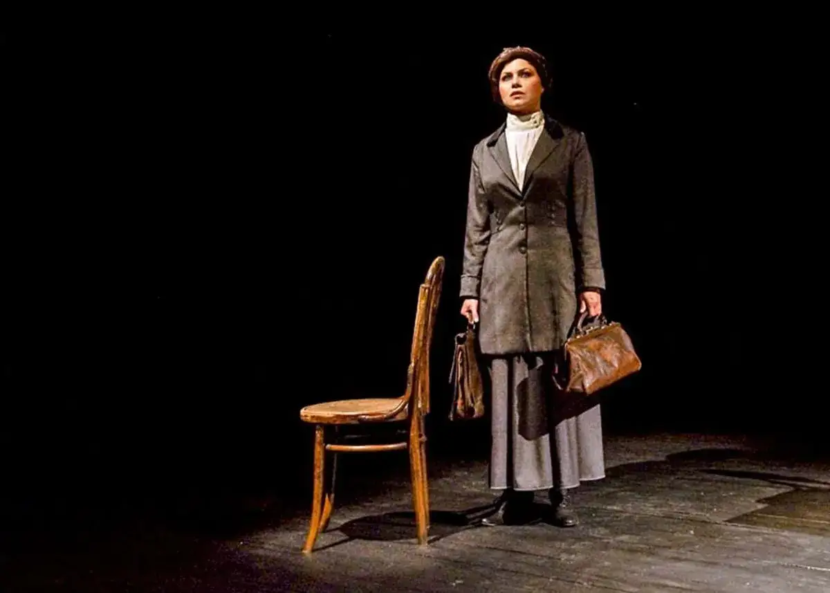 Sylwia Janowicz-Dobrowolska w historycznym kostiumie, z dwiema torbami, stoi obok drewnianego krzesła na ciemnej scenie.