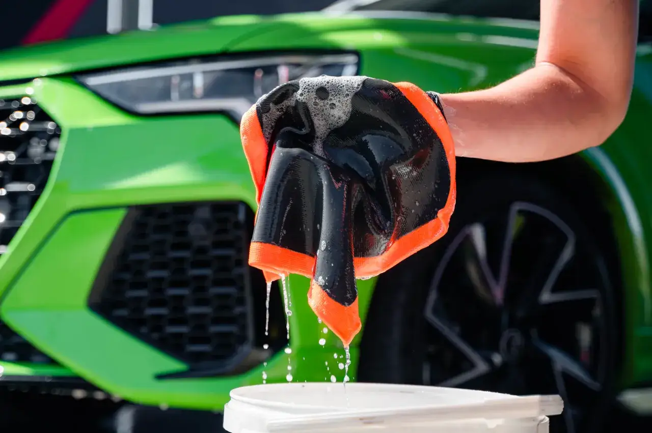 Piana spływa z rękawicy podczas czyszczenia lakieru samochodowego. Zielony samoch&oacute;d w tle.
