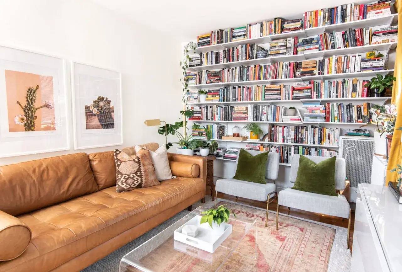 Przytulny salon z brązową kanapą, dwoma fotelami i regałem pełnym książek na tle białej ściany.
