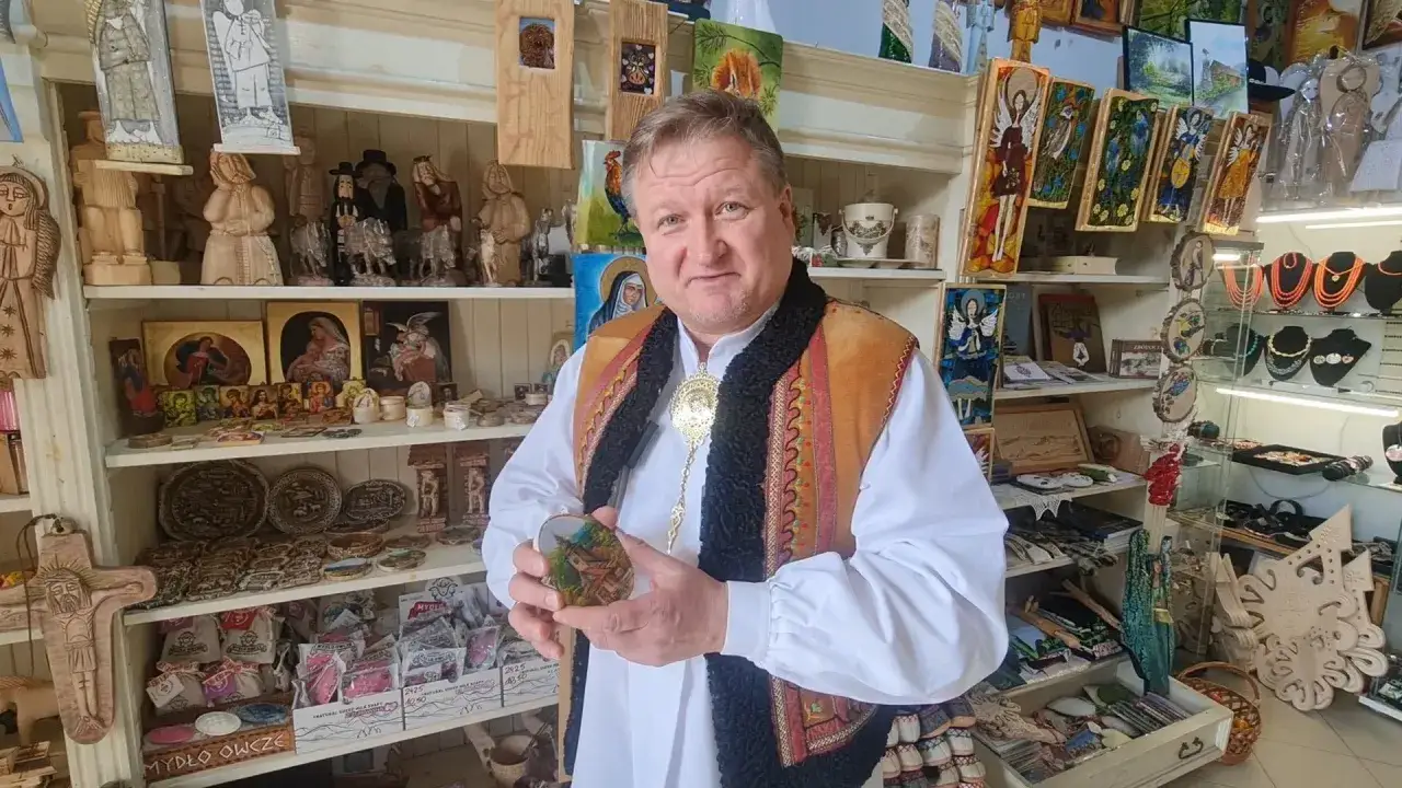 Mężczyzna w stroju ludowym trzyma ręcznie malowaną pamiątkę z wakacji. W tle półki pełne rękodzieła i lokalnych wyrobów.