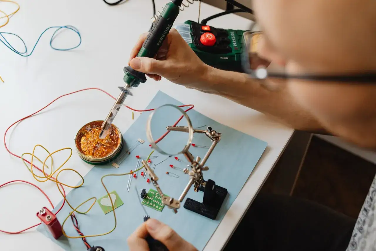Technik elektryk lutuje elementy elektroniczne, tworząc obwód. Na stole leżą przewody, diody LED, bateria i lupa.