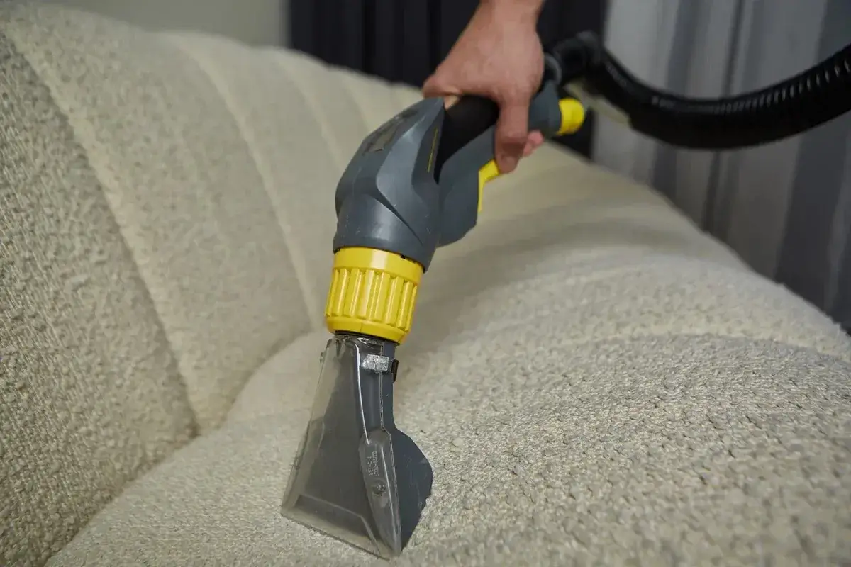 Czyszczenie kanapy materiałowej: ręka trzyma końc&oacute;wkę odkurzacza piorącego, kt&oacute;ra zbliża się do jasnej tkaniny mebla.